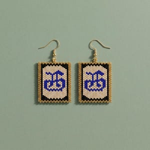 May include: A pair of beaded earrings with a rectangular design. The earrings feature a blue and white pattern within a black border, framed by gold-colored beads. The earrings hang from gold-colored hooks.