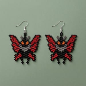 May include: A pair of beaded butterfly earrings. The earrings have a black body, red wings with black accents, and grey details. The butterfly's eyes are orange. The earrings hang from silver-coloured hooks.
