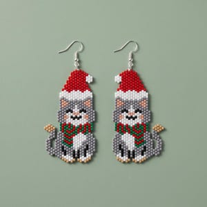 Puede incluir: Un par de pendientes de cuentas que muestran un gato gris con un gorro de Papá Noel rojo y blanco y una bufanda a rayas rojas y verdes. El gato está representado en estilo pixelado con cuentas pequeñas y cuelga de un gancho de metal plateado.