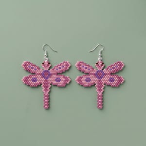 May include: A pair of dragonfly earrings. Each earring is crafted from small, pink and purple beads, forming a dragonfly shape. The wings are a lighter pink with darker purple accents. The earrings hang from silver-colored hooks.