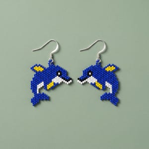 Könnte beinhalten: Ein Paar Ohrhänger mit delfinförmigen Anhängern aus kleinen blauen, weißen, gelben und schwarzen Perlen. Die Delfinfiguren haben ein pixeliges Aussehen und sind an silberfarbenen Metall-Ohrhaken befestigt.