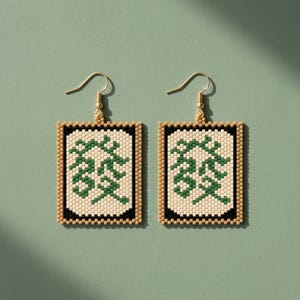 May include: A pair of rectangular beaded earrings. Each earring features a cream-colored center with green Chinese characters, framed by black and tan beads. Gold-colored ear wires complete the design. The earrings are displayed against a muted green background.