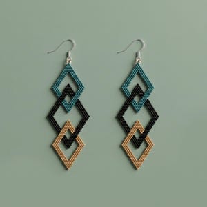 Pode incluir: Um par de brincos pendentes com três armações em forma de diamante entrelaçadas feitas de pequenas contas. O diamante superior é azul-petróleo, o do meio é preto e o inferior é bege. Cada brinco pende de um gancho de metal prateado.