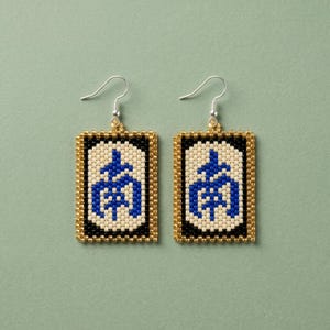 May include: A pair of beaded earrings with a rectangular design. Each earring features a blue symbol on a cream background, framed by black and gold beads. The earrings have silver-colored ear wires.