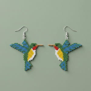 Pode incluir: Um par de brincos de contas em forma de beija-flor. Os brincos apresentam um design azul, verde, amarelo, vermelho e branco. As asas do beija-flor são azuis e o corpo é verde, amarelo e branco. Os brincos penduram-se em ganchos prateados.