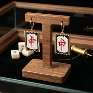 May include: Gold-toned drop earrings featuring a rectangular design with a white center, a red symbol, and black and gold borders. The earrings are displayed on a wooden stand, with Mahjong tiles in the background.