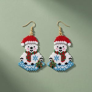 May include: A pair of beaded dangle earrings featuring a white polar bear wearing a red Santa hat and a green and red striped scarf. The bear is sitting on a light blue textured base with a small green gift box and a blue snowflake design.