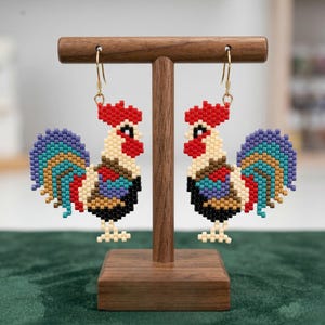 May include: A pair of beaded rooster earrings. The earrings feature a red comb, a cream-colored body, and a colorful tail with blue, green, and gold beads. The rooster's chest is black, and the earrings hang from gold-colored hooks.