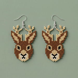 May include: A pair of beaded earrings shaped like deer heads, crafted from small square beads in shades of brown, cream, and black. The design features prominent antlers and a detailed facial structure, suspended from silver-toned metal earring hooks.