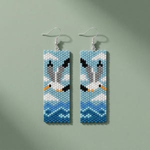 May include: Rectangular beaded earrings depicting seagulls. The earrings have a blue background with white clouds and a blue ocean. The seagulls are grey and black with orange beaks. The earrings have silver hooks.