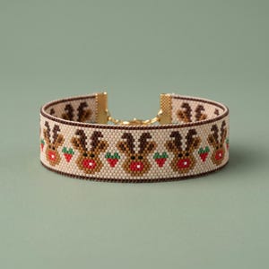May include: A beaded bracelet featuring a repeating pattern of brown reindeer heads with red noses and small green and red strawberries on a cream background, finished with gold-toned metal end caps and a lobster claw clasp closure.