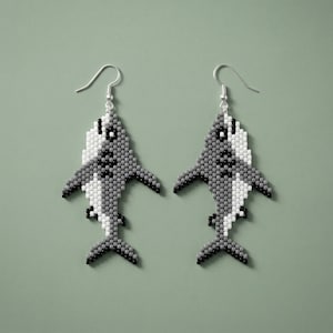 May include: A pair of beaded shark earrings. The earrings are gray, white, and black, with a fish hook closure. The sharks have a detailed design, with fins and a tail. The earrings are on a green background.