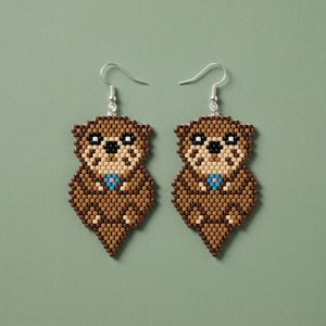 May include: A pair of beaded otter earrings. The earrings are brown, tan, and black, with a small blue flower detail. The earrings have silver-colored fish hook ear wires and are set against a green background.
