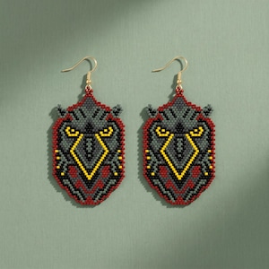 May include: A pair of dangle earrings crafted from small beads in a shield shape. The design features a stylized bird face in shades of dark gray, black, and red, with bright yellow accents forming the eyes and beak, suspended from gold-tone hooks.