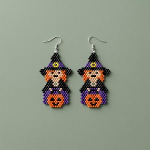 May include: A pair of beaded earrings featuring a witch character wearing a black and purple hat with a yellow accent, orange hair, and a black dress, holding a carved orange pumpkin. The earrings are constructed with small square beads.