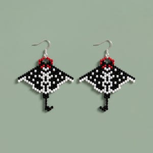 Könnte beinhalten: Ein Paar Ohrhänger aus kleinen Perlen in Form eines Mantarochens. Das Design zeigt ein geometrisches Muster in Schwarz und Weiß mit einem roten Akzent an der Oberseite, befestigt an silberfarbenen Metallhaken für Ohrlöcher.