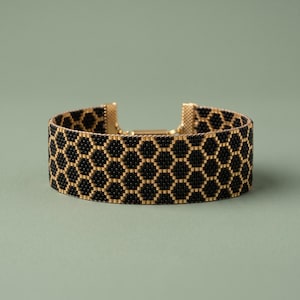 May include: A wide cuff bracelet crafted from small, uniform beads arranged in a repeating black and gold honeycomb pattern. The bracelet features a metallic gold clasp mechanism at the center, connecting the two ends of the beaded band.