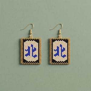 May include: A pair of rectangular beaded earrings. Each earring features a gold-colored border, surrounding a black and off-white background. A blue symbol is centered on the background. The earrings have gold-colored hooks.