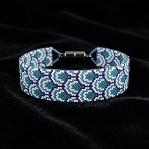 May include: A beaded bracelet featuring a repeating pattern of teal, white, and navy blue arcs. The bracelet has a magnetic clasp and is displayed on a black surface. The beaded design creates a textured appearance.