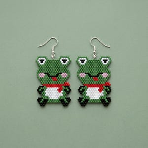 May include: A pair of handmade frog earrings. Each earring features a green beaded frog with white eyes, pink cheeks, a red scarf, and a white belly. The earrings have silver-colored fish hook ear wires. The background is a solid green color.