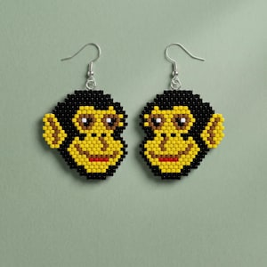 May include: A pair of beaded earrings shaped like monkey faces. The design features black, yellow, brown, white, and red beads arranged in a pixelated style. Each earring hangs from a silver-toned metal hook, creating a dangling accessory.