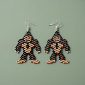 May include: A pair of dangle earrings crafted from small square beads in a pixelated design of a creature. The figure is composed of dark brown, light brown, and black beads, featuring a wide torso, long arms, and legs, suspended from silver hooks.