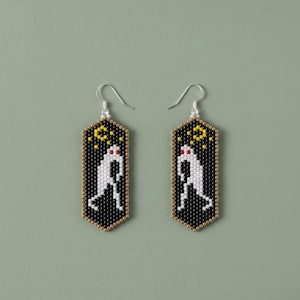 May include: A pair of hexagonal beaded earrings featuring a black background with a white ghost figure, red eyes, and a yellow crescent moon and stars. The earrings are constructed from small glass beads and hang from silver-toned metal ear wires.
