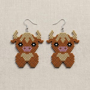 May include: A pair of handmade beaded earrings featuring a cartoon bull design. The earrings are made with small, colorful beads in shades of brown, tan, and pink, with silver-colored ear wires. The bull has horns, eyes, and a pink mouth.