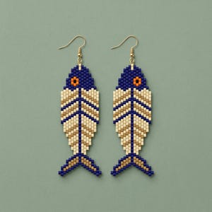 Op de afbeelding: Een paar kralen oorbellen in de vorm van een vis. De oorbellen hebben een blauwe kop, een oranje oog en een crème en gouden lichaam. De oorbellen hangen aan goudkleurige oorhaken. De achtergrond is effen blauwgroen.