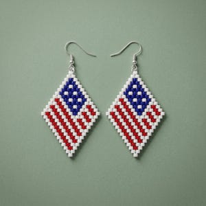 May include: A pair of diamond-shaped dangle earrings crafted from small square beads. The design features a pattern of red and white horizontal stripes with a blue section containing white dots, mimicking the American flag, on silver-tone hooks.
