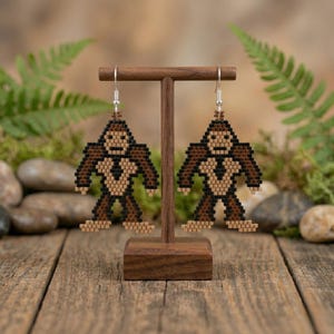 Puede incluir: Un par de pendientes con forma de Bigfoot. Los pendientes están hechos de pequeñas cuentas marrones, negras y beige. Los pendientes cuelgan de un expositor de madera. El fondo incluye vegetación y una superficie de madera.