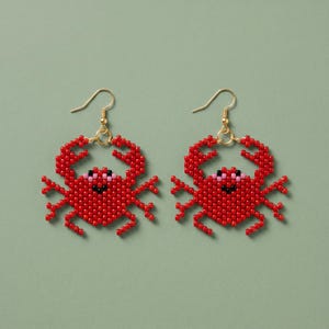 May include: A pair of red beaded crab earrings with gold-colored hooks. The crab earrings have black eyes and pink accents. The earrings are set against a light green background.