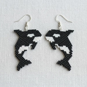 May include: A pair of beaded earrings featuring a black and white orca design. The earrings are made with small beads and have silver-colored ear wires. The orcas are depicted in a stylized, cartoonish manner.