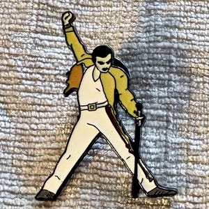 May include: A metal enamel pin depicting a performer in a yellow jacket, white shirt, and white trousers, standing with one arm raised and holding a microphone stand. The figure has dark hair and is rendered in a graphic, outlined style.
