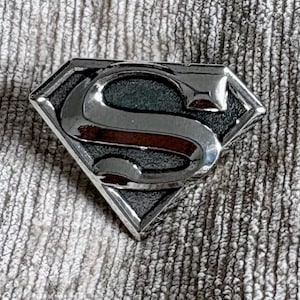 May include: A metallic Superman logo pin featuring a polished silver-tone finish on the raised letter S and the outer diamond-shaped border, contrasted against a recessed, dark, textured background within the shield-shaped emblem.