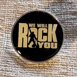 May include: A circular black pin featuring the gold-coloured text WE WILL ROCK YOU. The letter O in the word ROCK is replaced by a silhouette of a person holding a microphone stand in a dynamic pose, set against a glossy, smooth surface finish.