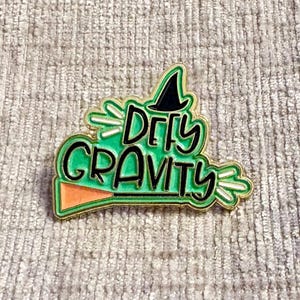 May include: A gold-rimmed enamel pin featuring a bright green background with the black text "DEFY GRAVITY". The design includes a black witch hat at the top and an orange broomstick at the bottom, accented with white motion lines on the sides.
