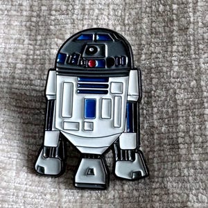 May include: A metal enamel pin shaped like the droid R2-D2, featuring a white, blue, and dark grey colour scheme. The design includes a domed head with a red circular detail, a central blue rectangular panel, and various geometric white and blue accents.