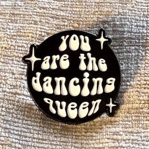 May include: A circular black enamel pin featuring the white text "you are the dancing queen" in a rounded, bubbly font. The design includes three small, four-pointed star accents positioned around the text, creating a high-contrast graphic.