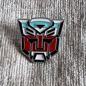 May include: A metal enamel pin featuring the Autobot insignia from Transformers. The design is shaped like a stylised robot face with a metallic silver border, filled with sections of white, bright red, and a light turquoise blue enamel.