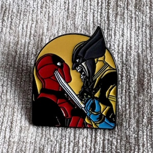 May include: A metal enamel pin featuring a graphic illustration of two comic book characters facing each other. One character is red and black, while the other is yellow, black, and blue. They are set against a circular yellow background.