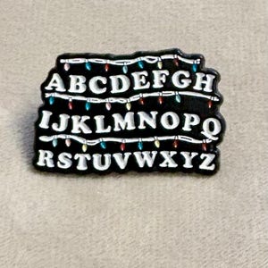 May include: A black enamel pin featuring three horizontal rows of white capital letters from A to Z. Between each row of letters, there is a string of small, colourful decorative light bulbs in shades of red, blue, green, and yellow.