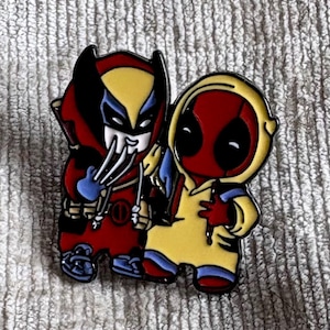 May include: A metal enamel pin featuring two cartoon characters standing side by side. The left character wears a red and yellow mask with silver claws, while the right character wears a red mask and a yellow hooded jacket with black accents.