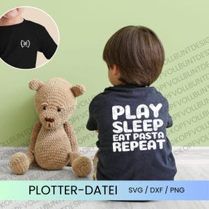 May include: A dark navy blue t-shirt featuring white text on the back that reads PLAY SLEEP EAT PASTA REPEAT. A circular inset shows the front of the shirt with a small white bow tie pasta graphic on the chest. A tan crocheted teddy bear is visible.