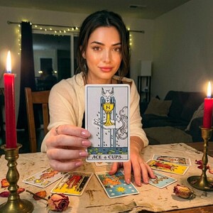 K&ouml;nnte beinhalten: Eine Frau h&auml;lt eine Tarotkarte mit dem Text "ACE of CUPS". Die Karte zeigt einen goldenen Kelch und eine Taube. Mehrere andere Tarotkarten liegen auf einem Tisch, zusammen mit zwei brennenden roten Kerzen in Messinghaltern.