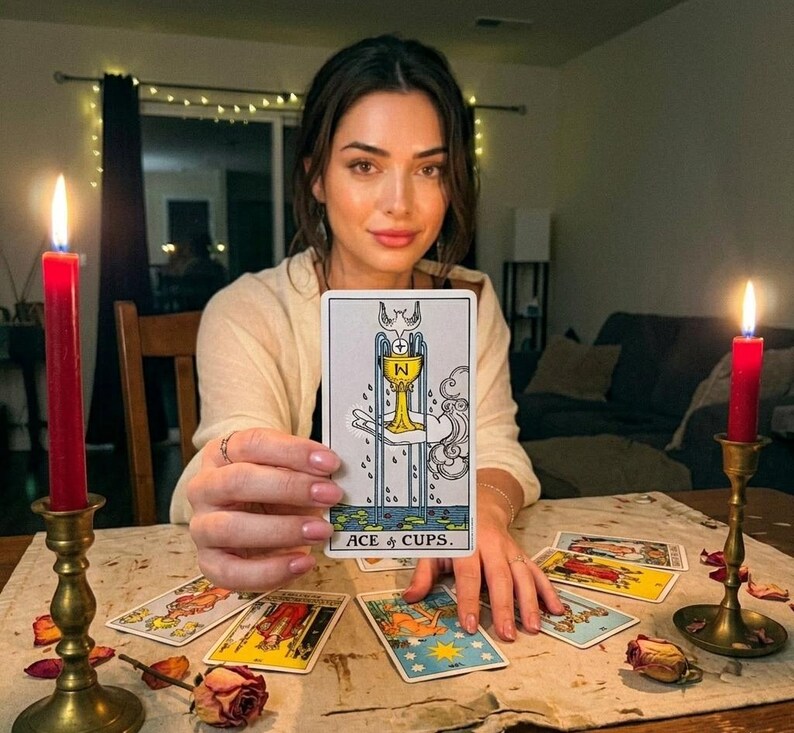 K&ouml;nnte beinhalten: Eine Frau h&auml;lt eine Tarotkarte mit dem Text "ACE of CUPS". Die Karte zeigt einen goldenen Kelch und eine Taube. Mehrere andere Tarotkarten liegen auf einem Tisch, zusammen mit zwei brennenden roten Kerzen in Messinghaltern.