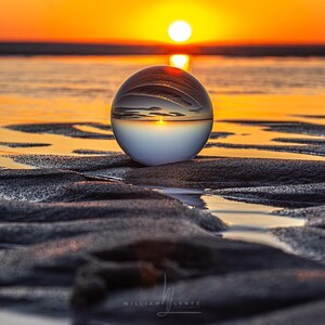 Può includere: Una sfera di vetro trasparente riflette un tramonto vibrante su una spiaggia sabbiosa. La sfera è posizionata su sabbia scura e strutturata, con il bagliore del sole specchiato al suo interno. Il cielo passa dall'arancione al giallo, creando una scena naturale serena.