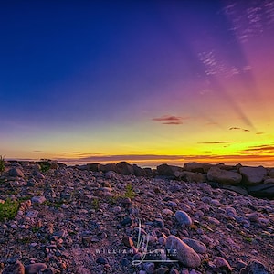 Op de afbeelding: Een kustlandschap met een wit en blauw geschilderde vuurtoren onder een kleurrijke zonsondergang. De voorgrond is een rotsachtige kust. De lucht gaat over van blauw naar paars en geel, met lichtstralen.