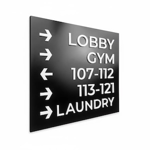 May include: A rectangular black directional sign with white raised lettering and arrows. The text reads LOBBY, GYM, 107-112, 113-121, and LAUNDRY, each accompanied by a white arrow pointing to the left or right to indicate the direction for each location.