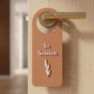 May include: A terracotta-colored door hanger featuring the white text In Session and a white botanical leaf graphic. The rectangular sign has a circular cutout at the top, allowing it to hang securely from a brass-toned metal door handle.
